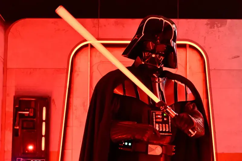 The image depicts a character in a dark, armored suit holding a glowing lightsaber, set against a red-lit background.