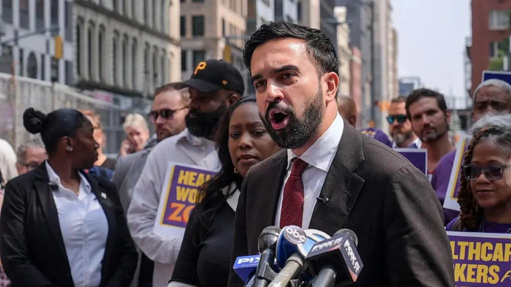 A man in a suit speaks into microphones at a press conference, surrounded by people holding signs advocating for healthcare workers.