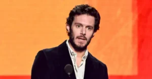The image shows a man speaking at a podium in front of a colorful background.