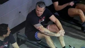 A man is seen in a gym setting, possibly during a workout or training session, with another individual observing.