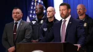A man in a suit stands at a podium, addressing an audience, with police officers in uniform standing behind him.