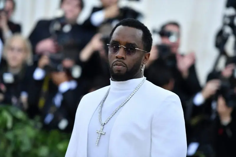 The image shows a person dressed in a white outfit with a large cross necklace, standing in front of a crowd of photographers.