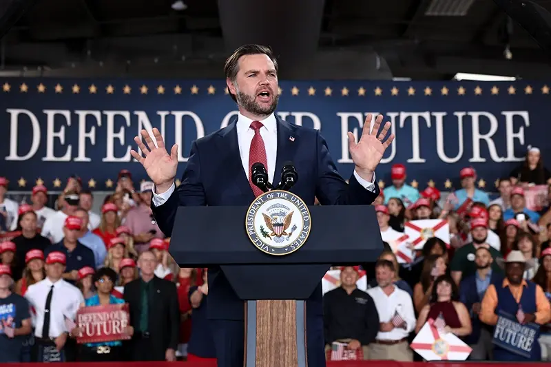 A man in a suit stands at a podium with a "Defend Our Future" sign in the background, addressing a crowd of supporters.