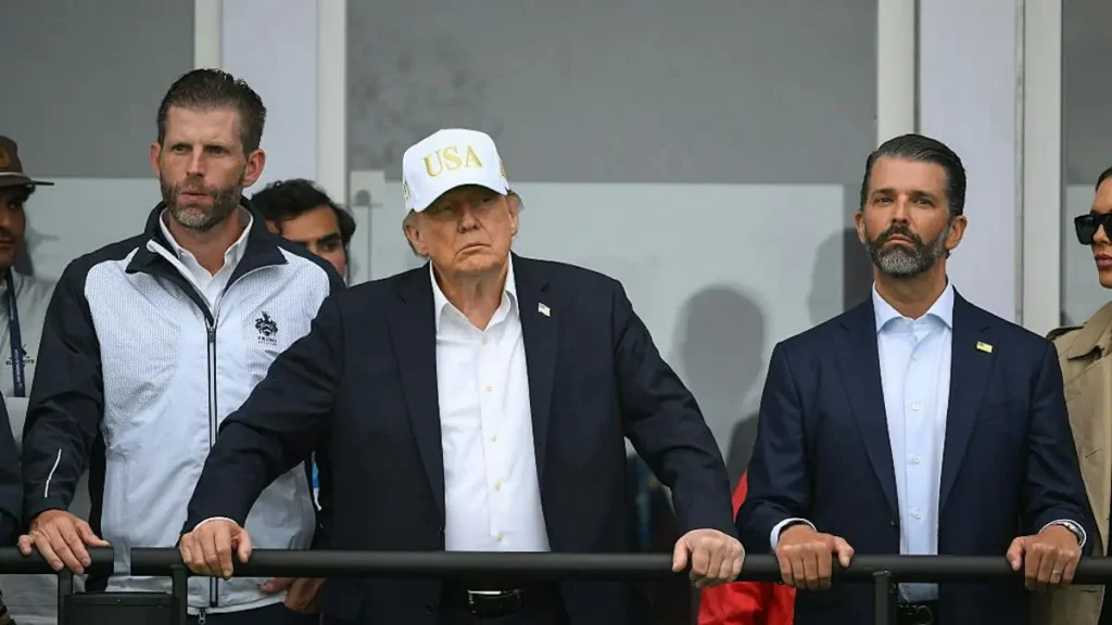 The image shows a group of individuals, including a man in a suit and a white cap with "USA" written on it, standing behind a railing.