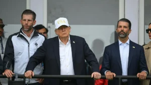 The image shows a group of individuals, including a man in a suit and a white cap with "USA" written on it, standing behind a railing.