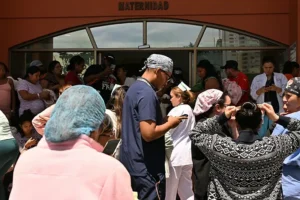 A group of people, including medical staff and patients, gather outside a hospital's maternity entrance.