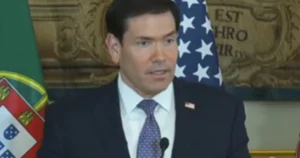 The image shows a man in a suit and tie speaking at a podium with flags of the United States and another country in the background.
