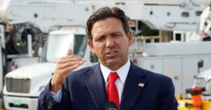 A man in a suit and red tie is speaking into a microphone in front of a large truck.