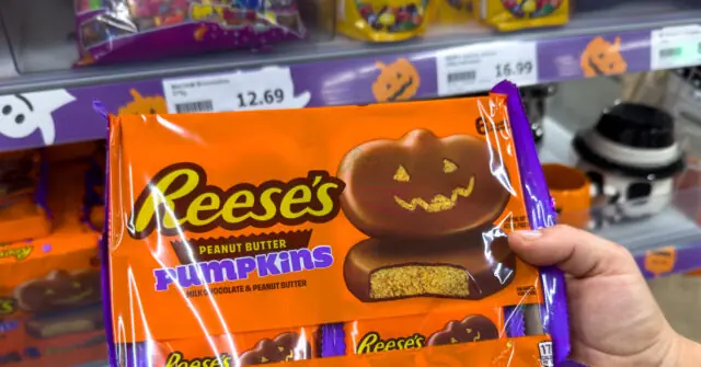 A hand holds a package of Reese's Peanut Butter Pumpkins, a Halloween-themed chocolate treat, in a store setting.
