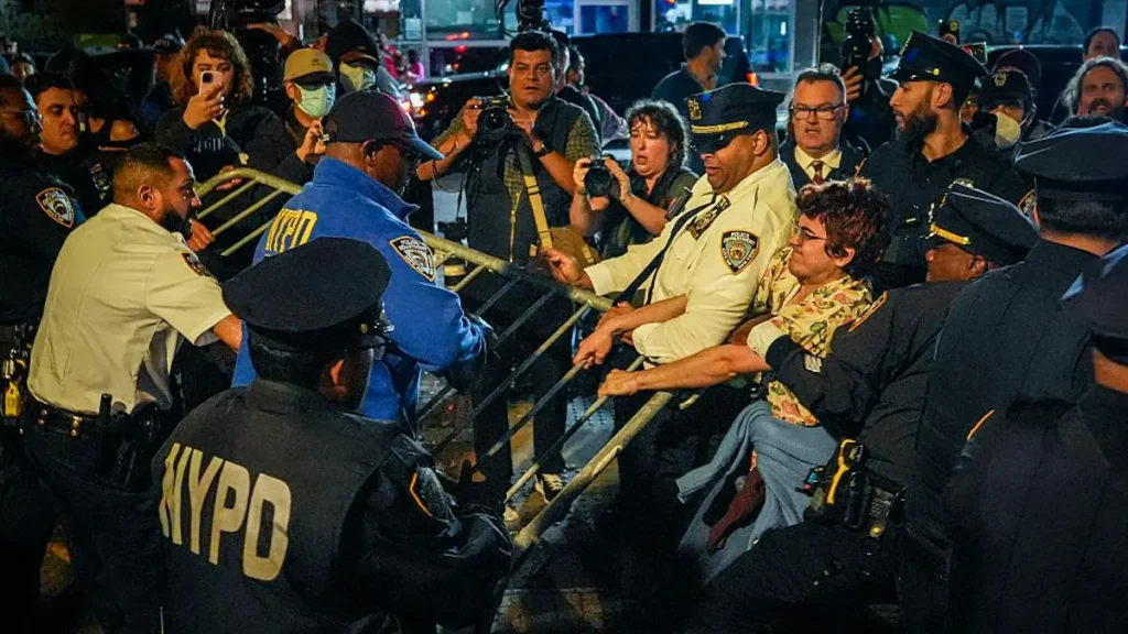A group of NYPD officers is seen managing a crowd of people, with some individuals holding onto a barricade, as media personnel capture the scene.