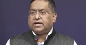 The image shows a man speaking into a microphone, dressed in formal attire, against a plain background.