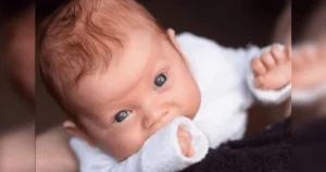 A baby wearing a white outfit is lying down and looking up with a curious expression.