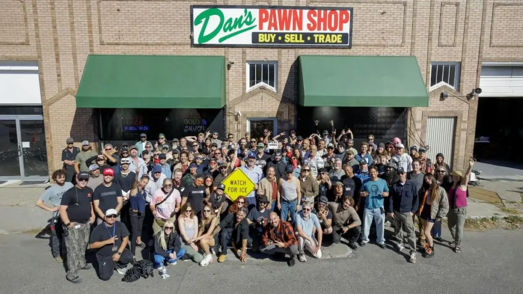 A large group of people poses for a photo in front of Dan's Pawn Shop, holding a sign that reads "WATCH FOR ICE."