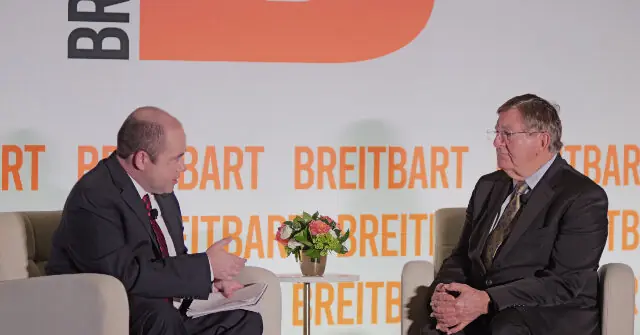Two men in suits are seated on a stage, engaged in a discussion at a Breitbard event.