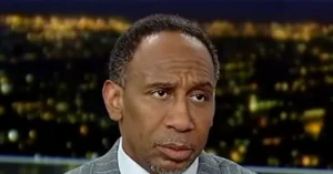 The image shows a man in a suit and tie, appearing to be on a news broadcast with a cityscape in the background.