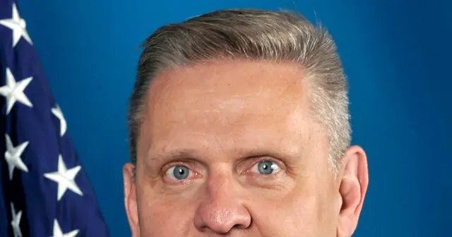 The image shows a close-up of a person with gray hair, set against a blue background with an American flag partially visible on the left.