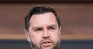 The image shows a close-up of a man with short dark hair and a beard, wearing a suit, against a blurred background.
