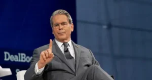 The image shows a man in a suit gesturing with his right hand while speaking at a formal event.