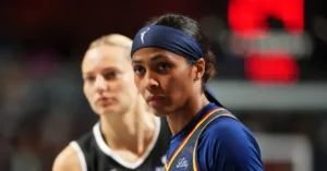 The image shows two basketball players in a game setting, with one player wearing a blue jersey and a headband, and the other in a black jersey.