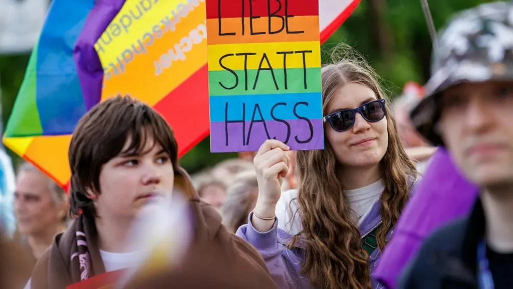 The photo captures a vibrant protest scene with a person holding a rainbow sign that reads "LIEBE STATT HASS," advocating for love over hate.