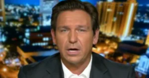 The image shows a news anchor in a studio setting with a cityscape background.