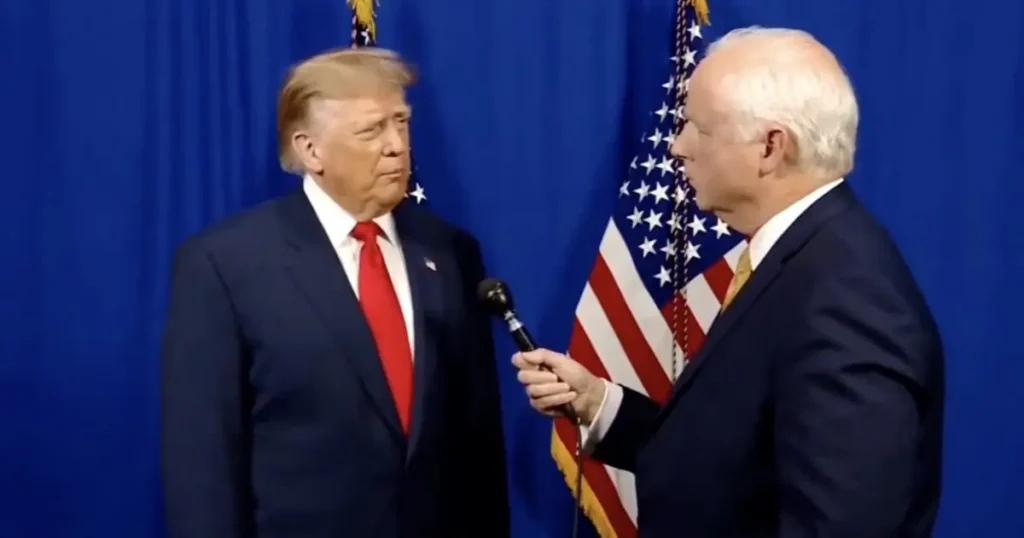 The image shows two men in suits standing in front of an American flag, with one holding a microphone, suggesting a formal interview or press event.