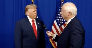 The image shows two men in suits standing in front of an American flag, with one holding a microphone, suggesting a formal interview or press event.