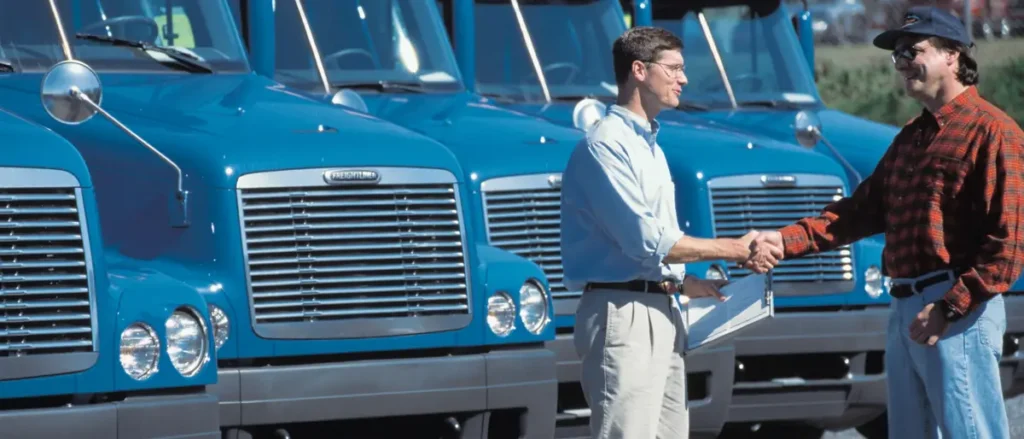 Two men shake hands in front of a row of blue trucks, possibly indicating a business deal or agreement.