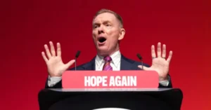 A man stands at a podium with a "HOPE AGAIN" sign, gesturing passionately with his hands raised.