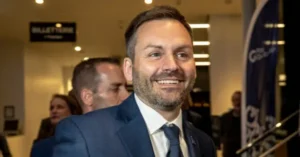 The image shows a man in a suit smiling in what appears to be a professional or formal setting, possibly at an event or conference.